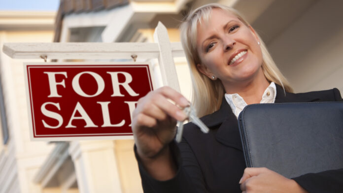 Female,Real,Estate,Agent,With,Keys,In,Front,Of,Sign